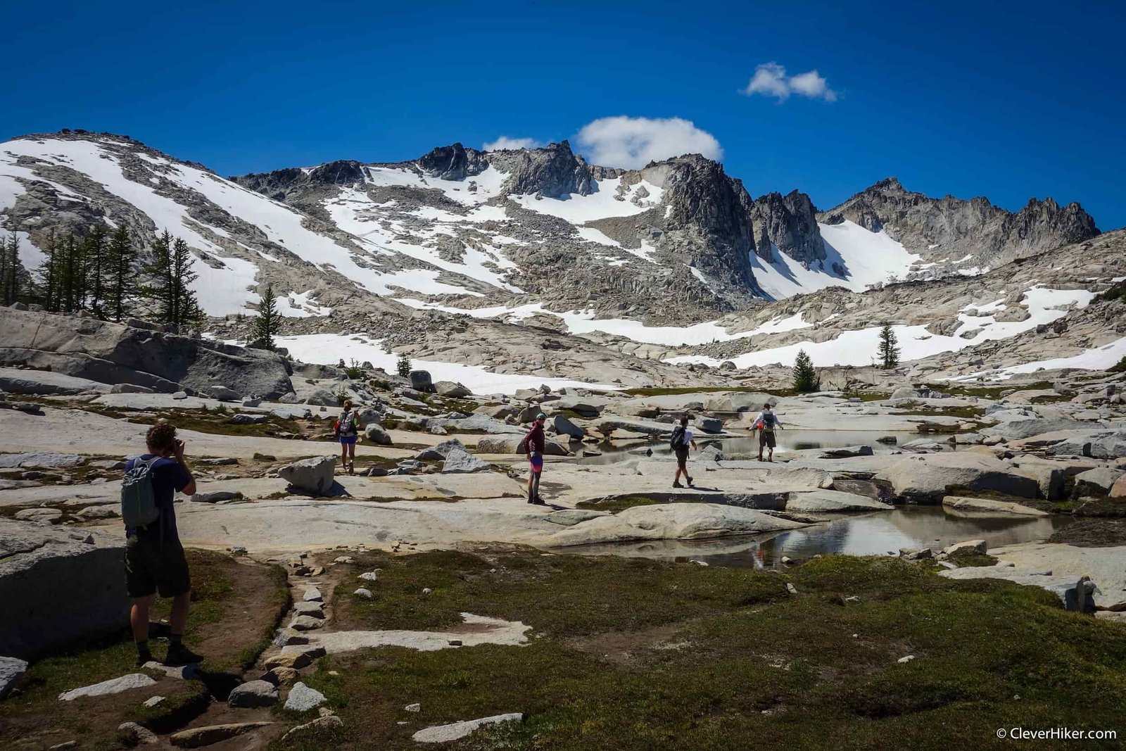 backpackers crossing granite slabs with snow lingering on the ridge.