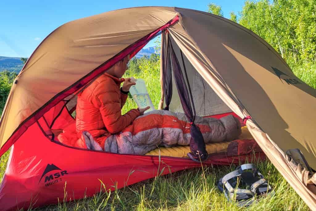 A hiker sips water in her tent while wrapped up in a down sleeping bag
