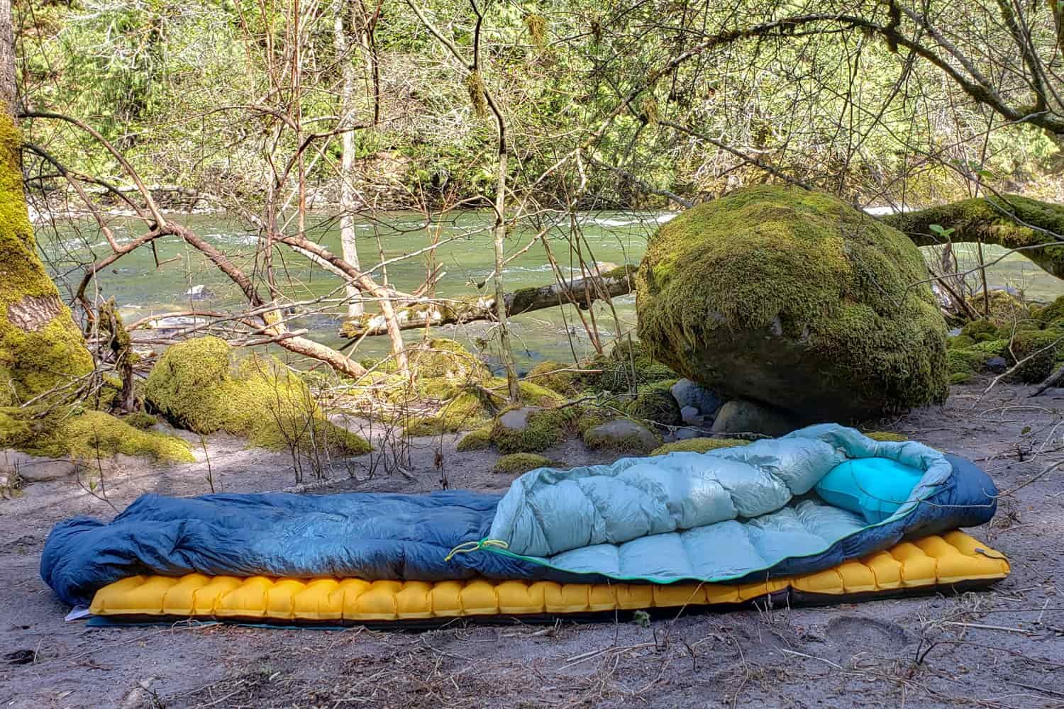 a sleeping bag and pad set up on a sandy beach bar next to a river in a forest.