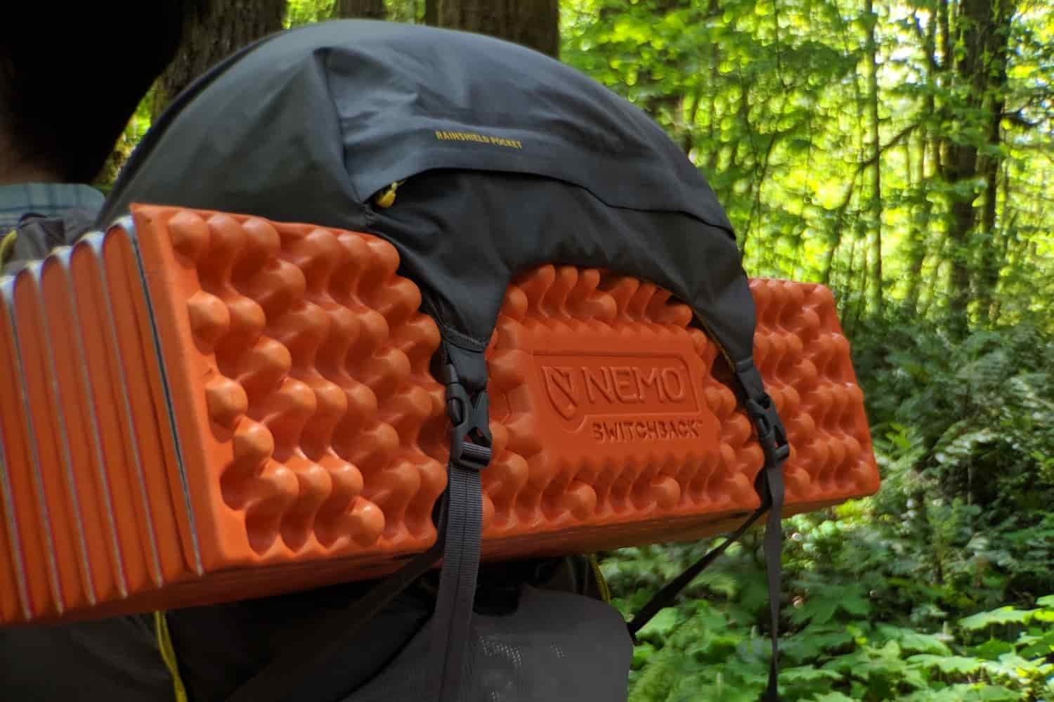 a backpacker's pack with the nemo switchback secured with two straps underneath the top "brain" pouch of the bag. They are in a brightly lit green hardwood forest.
