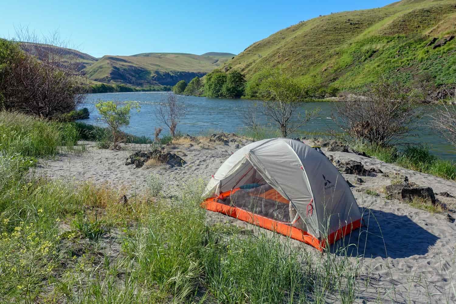 A tent is setup in a lush green valley near a lake.