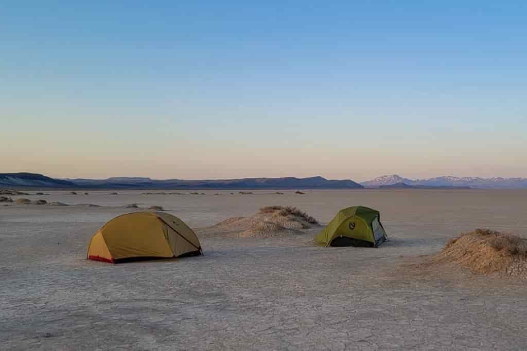 two tents pitched on a flat desert floor with purple mountains in the far distance 