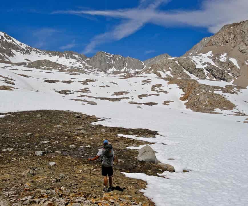 a man faces the camera with a big snow field behind him wearind trail runners
