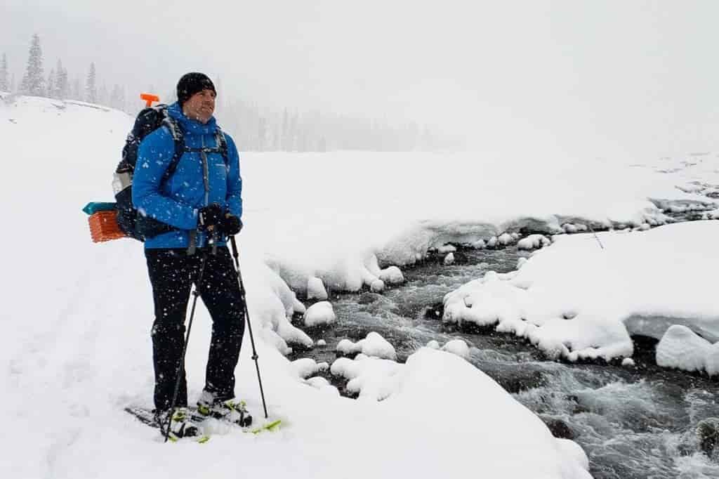 a backpacker stands next to a flowing creek in a winter snow covered wilderness. He is wearing snowshoes to move atop the snow