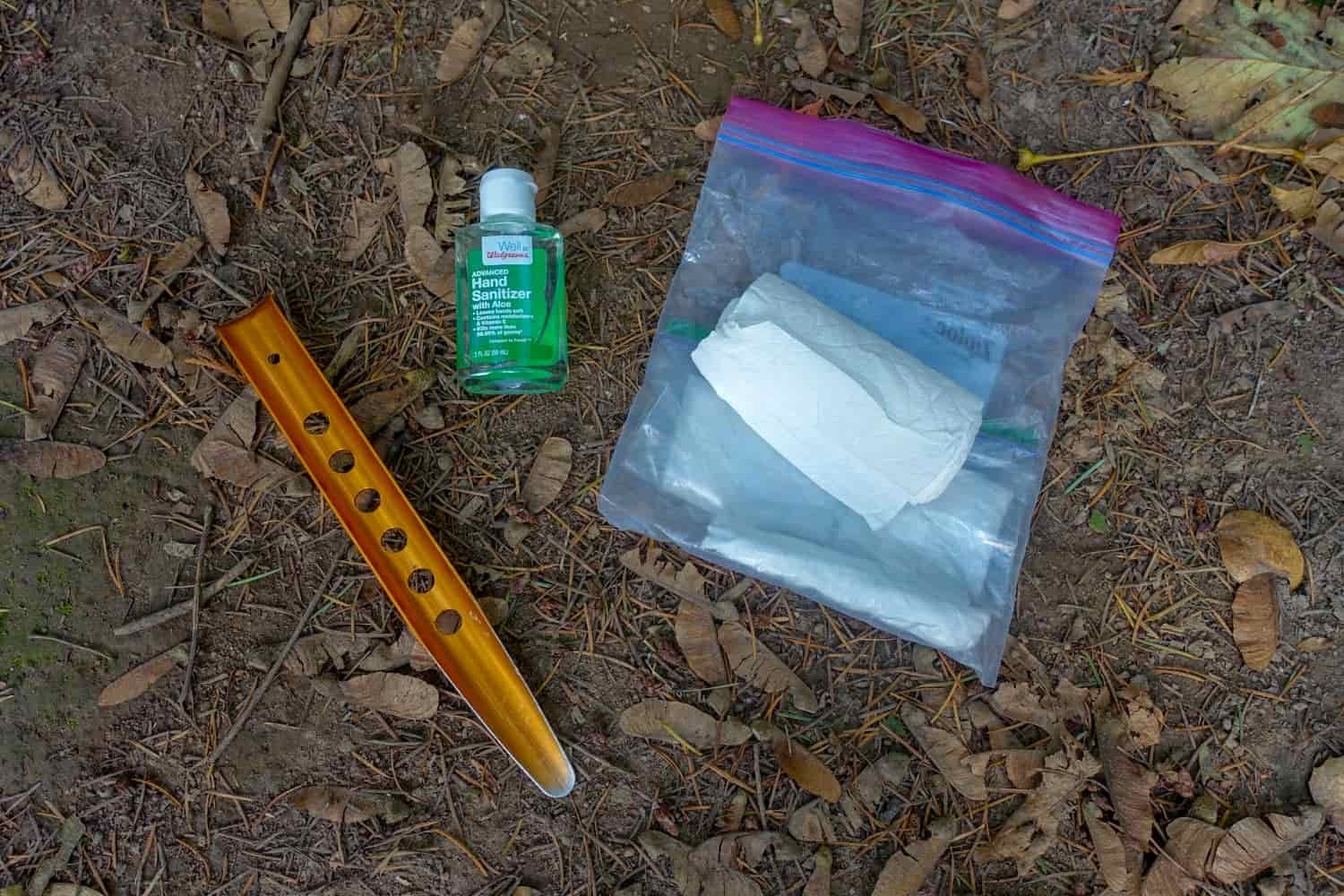 A snow stake, hand sanitizer, and Ziploc bag with toilet paper lay on the ground of a campsite