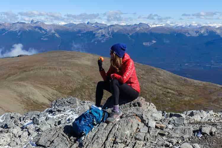 A female hiker sits on a summit eating a red apple in her red down puffy jacket