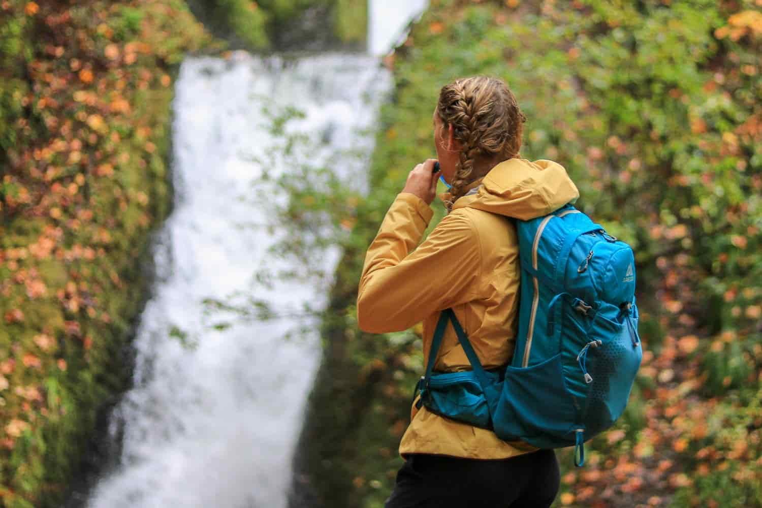 A female day hiker sips water from her hydration bladder while staring at a waterfall in front of her