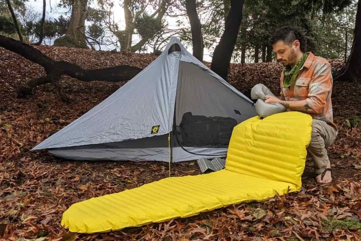 A hiker inflating a sleeping pad in front of the Six Moon Designs Lunar Solo Tent
