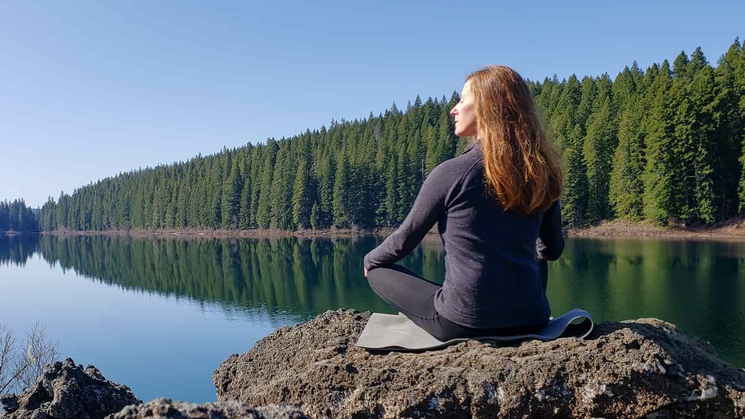 A woman sitting on the edge of a tree-lined lake in the Smartwool Classic Thermal Base Layers