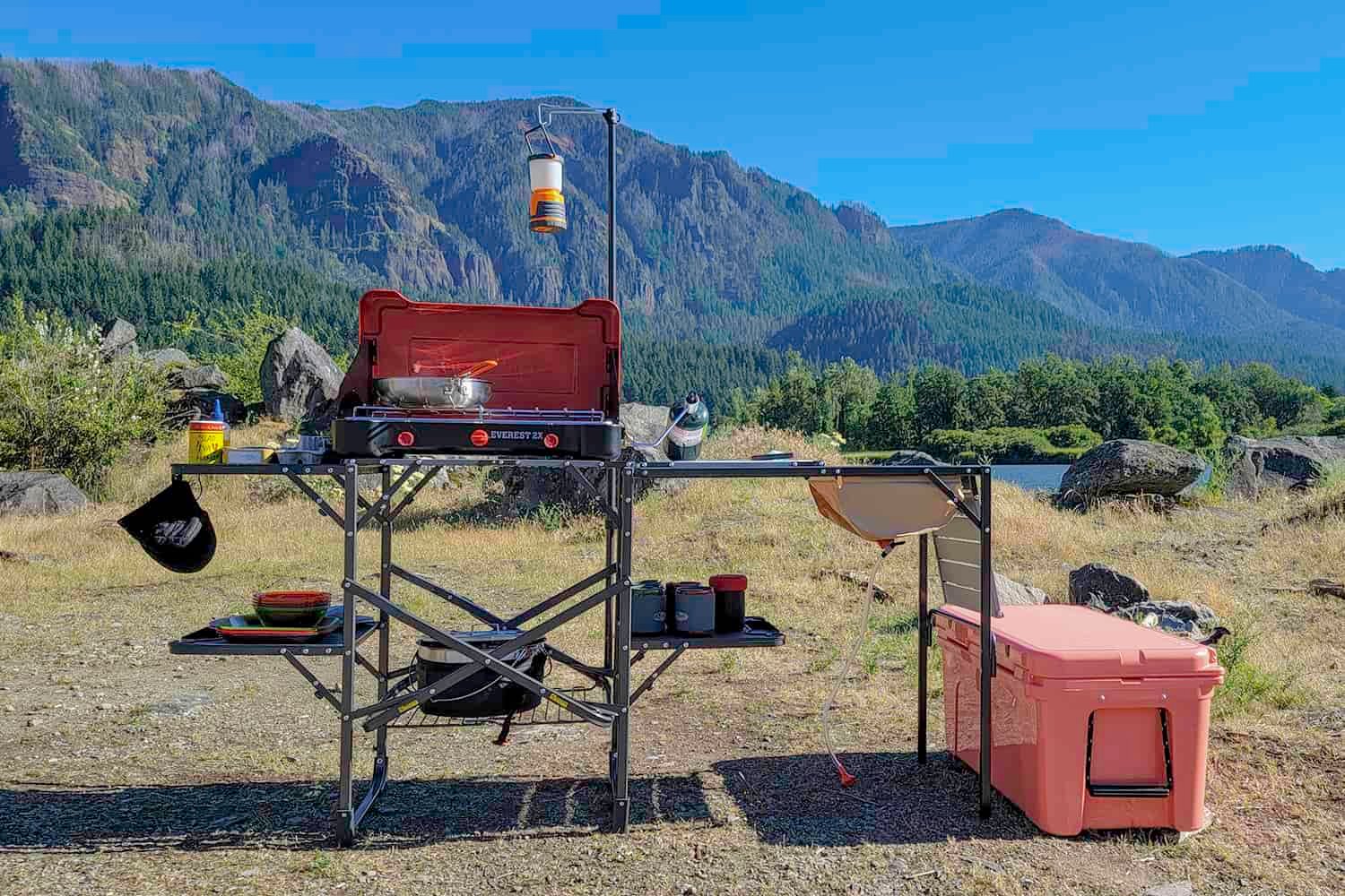 The GCI Outdoors Master Cook Station set up in a lakeside campsite with the Camp Chef Everest 2x stove, a cookset, a YETI Tundra Cooler, lantern, etc.