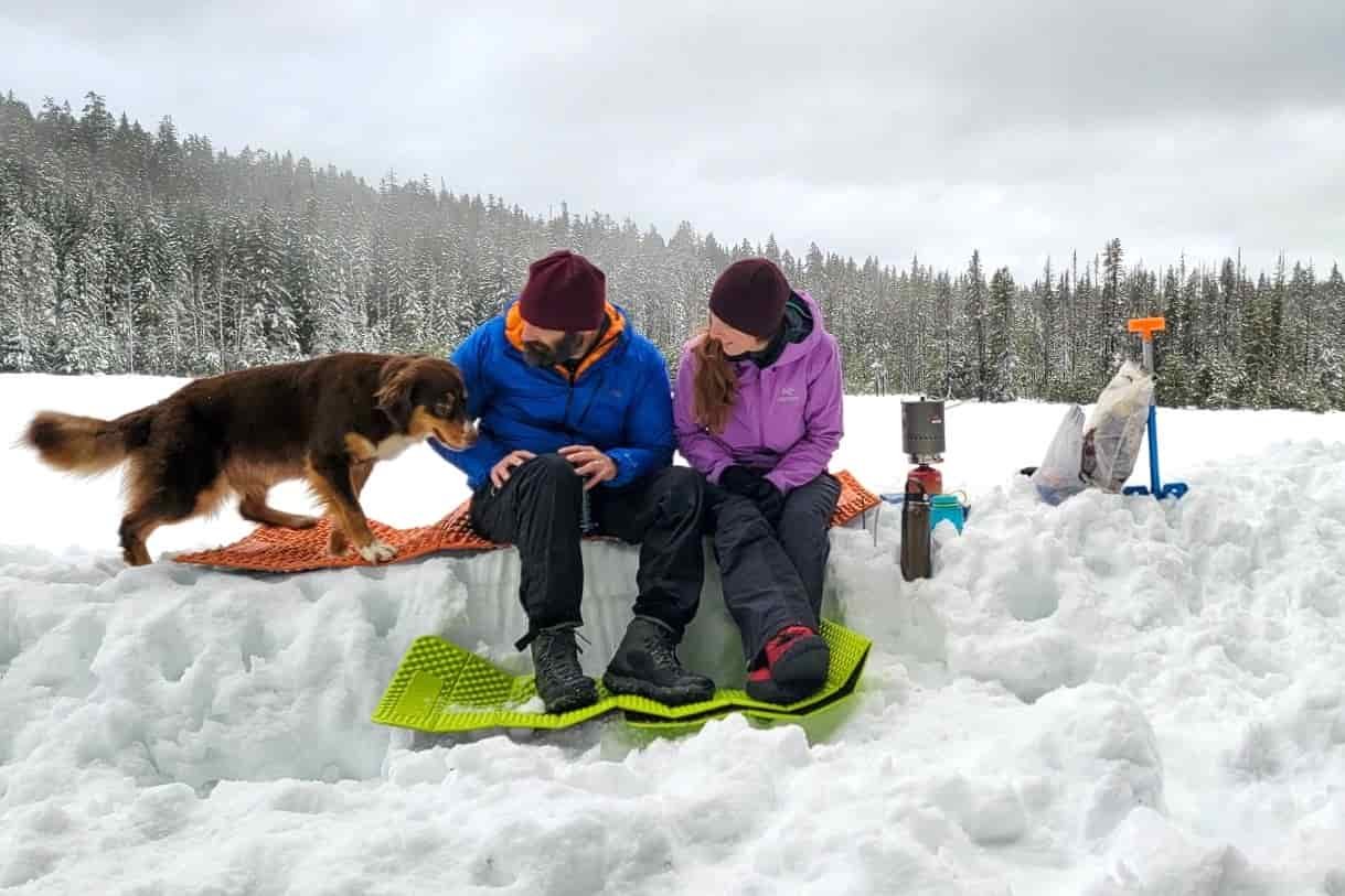 two people in a winter camp making food while sitting on a foam pad with their dog