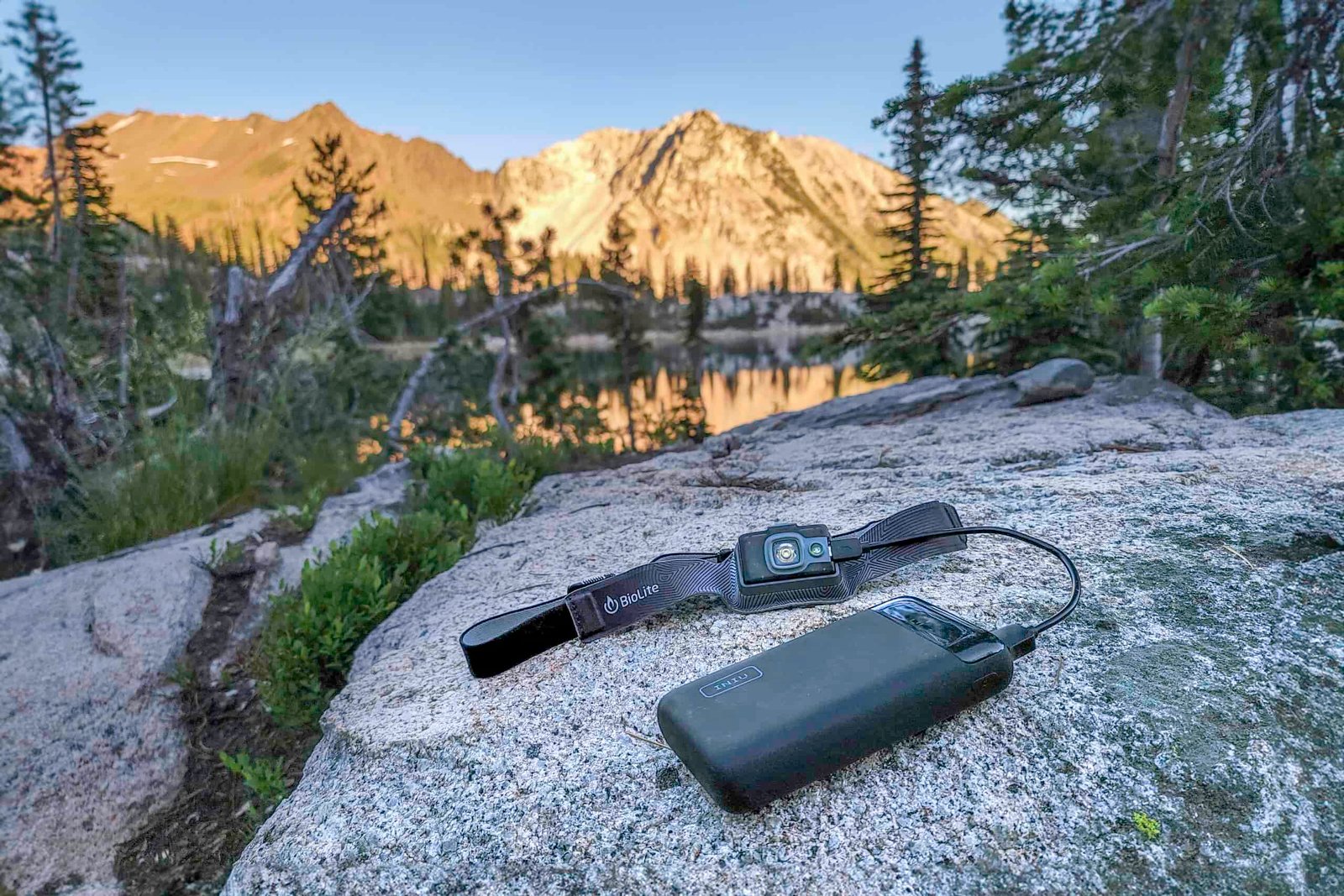 The Iniu 20000 PD power bank sitting on a granite slab while charging a rechargeable Biolite headlamp with the Wallowa Mountains at sunset in the background