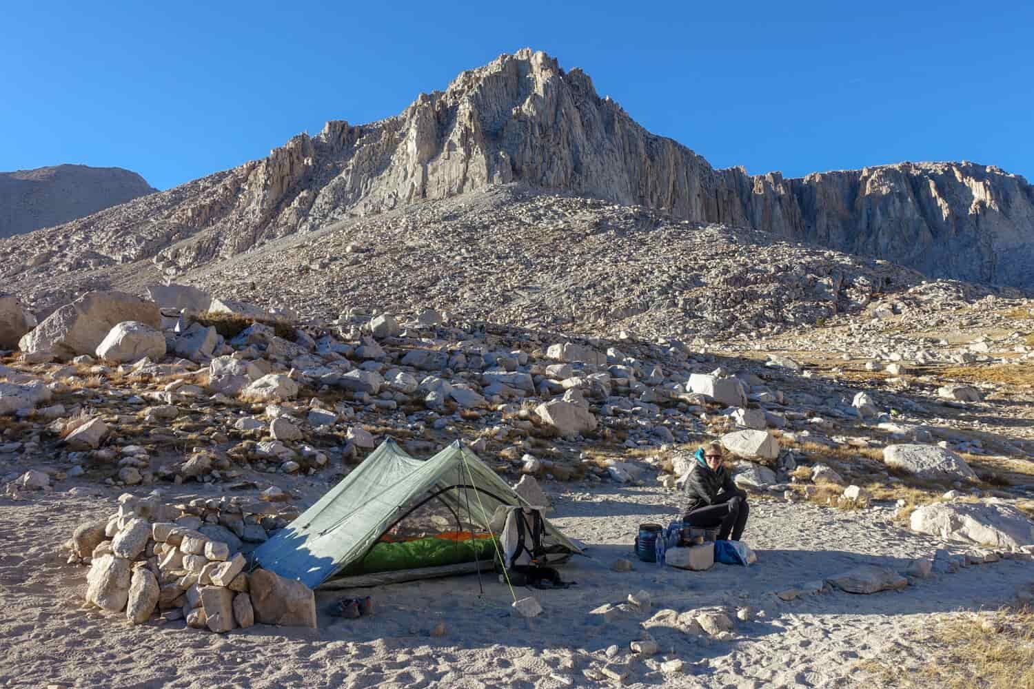 A woman sits near a pile of gear next to an ultralight tent set up above the treeline
