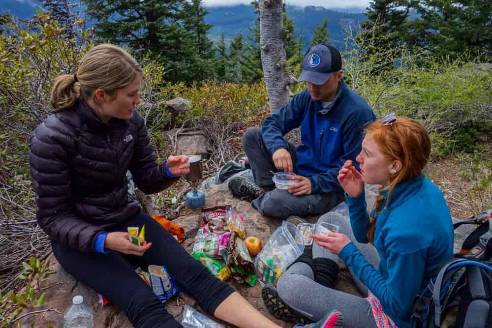 Small bags of food spread out on the ground as three backpackers eat lunch alongside the trail