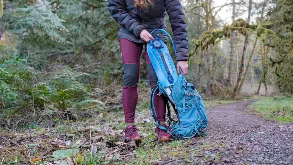 Waist-down view of a female hiker pulling the Gregory 3D Hydro hydration bladder out of her backpack along a trail