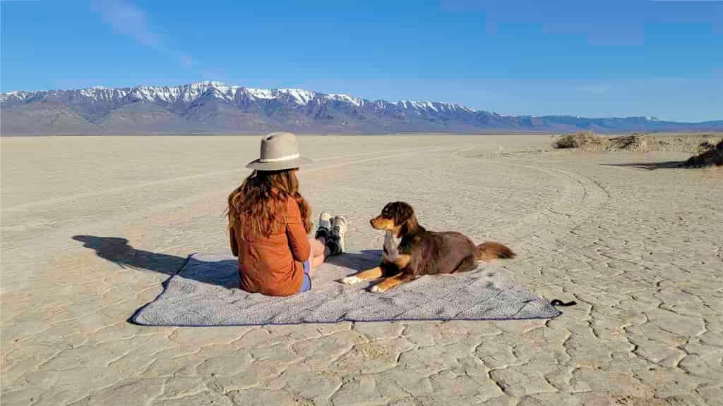A woman and a dog sitting on the Oceas Sherpa Fleece camping blanket in the Alvord Desert with the Steens Mountains in the background