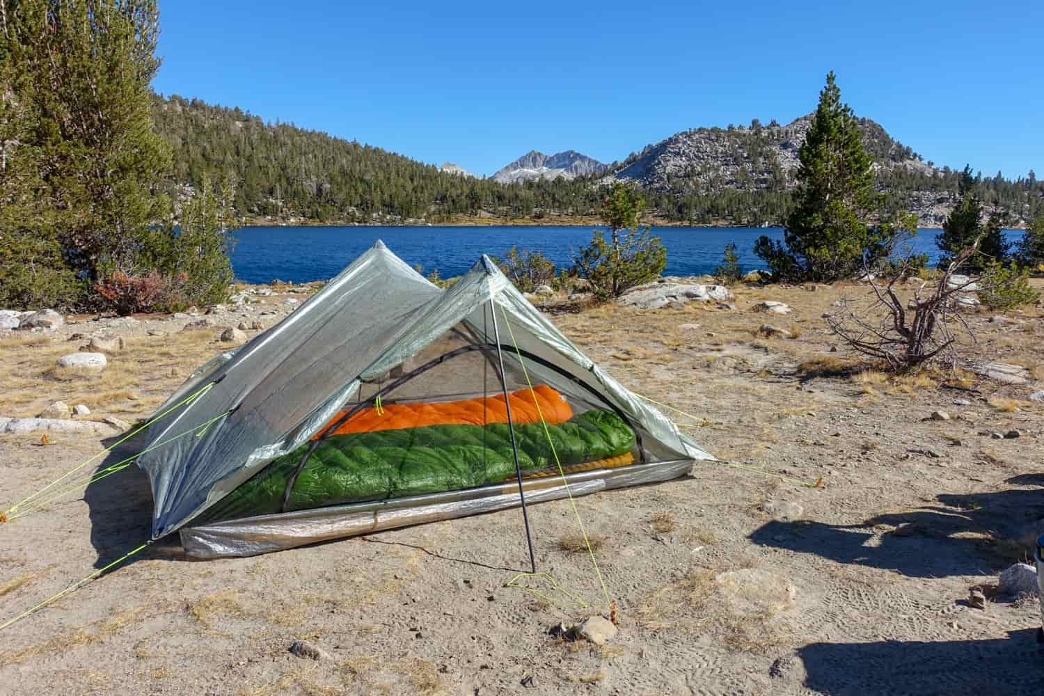 the zpacks triplex set up next to a large alpine lake on a cloudless sunny day with two sleeping pads and bags laid out inside