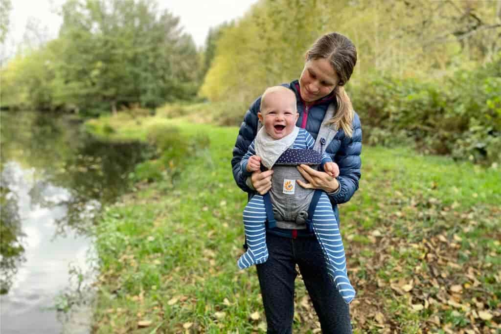 A mom looking down at her smiling baby in a front carrier while hiking near a pond