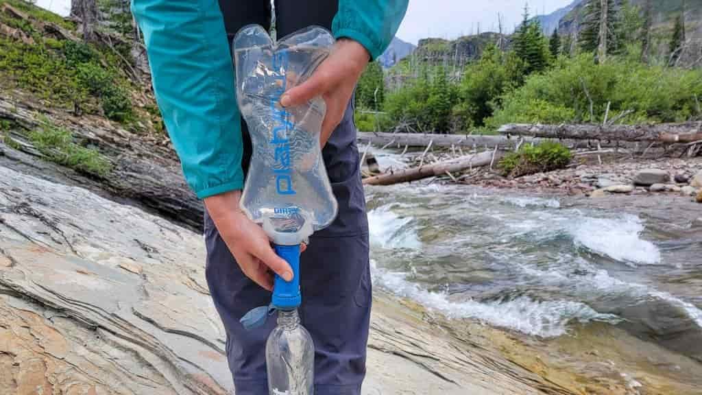 a backpacker stands near a stream with a water bottle on the ground between their feet for support while they squeeze a quickdraw water bladder through a filter.