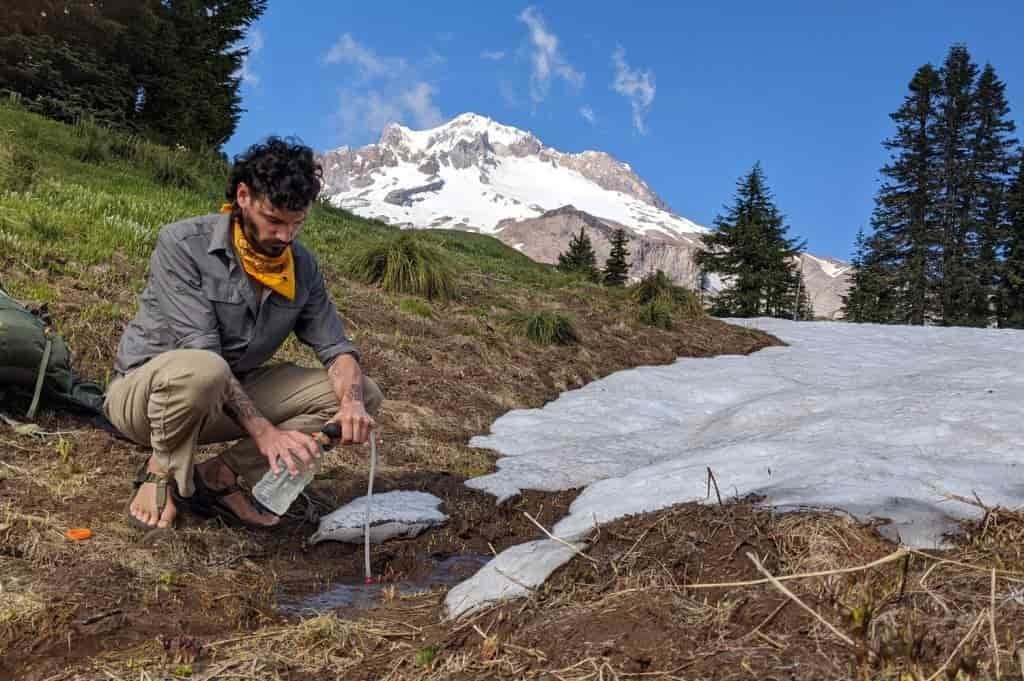 A hiker filters water downhill of a snow bank while wearing Bedford Cairn hiking sandals
