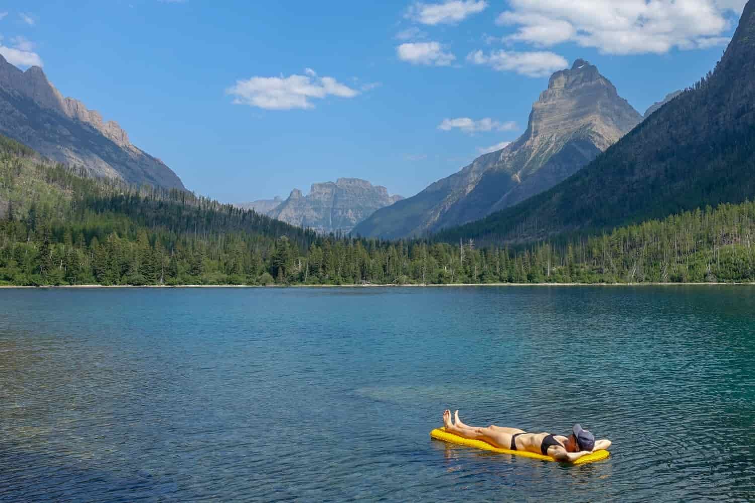 a hiker at a lake in glacier national park laying on their sleeping pad in the lake using it as a floaty