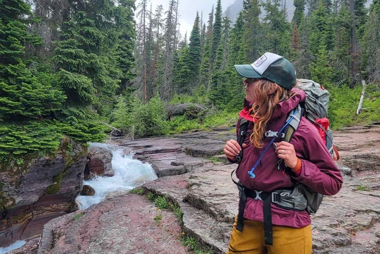 A female backpacker wearing the Rab Kinetic 2.0 rain jacket near a roaring creek in Glacier National Park