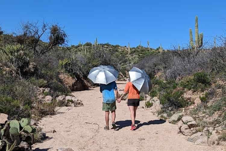 A couple holds hands as they walk down a desert wash holding umbrellas over their heads to shield themselves from the sun 