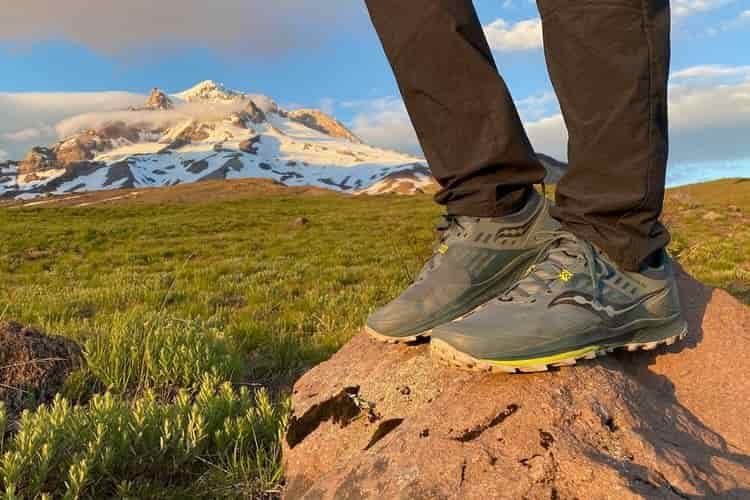 Close up of a hiker's trail running shoes as he stands on a rock with a snowy mountain in the background