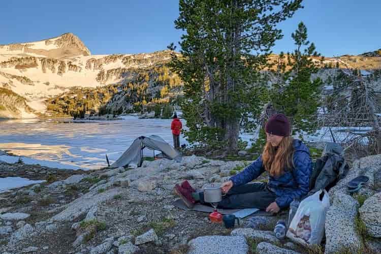 A woman works a camp stove at a campsite next to an icy alpine lake as another female backpacker looks over the frozen water