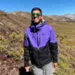 Against a summer mountain backdrop in the Colorado Rockies, a man in purple and black rain jacket and green sunglasses hikes towards camera while smiling