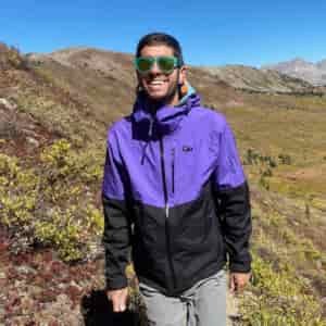 Against a summer mountain backdrop in the Colorado Rockies, a man in purple and black rain jacket and green sunglasses hikes towards camera while smiling