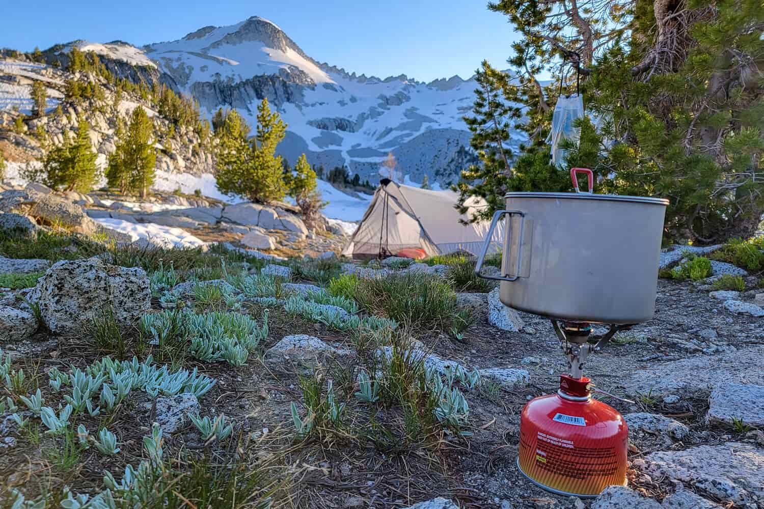 A backpacking stove boils water in a small pot with a tent and stunning mountain view in the background.