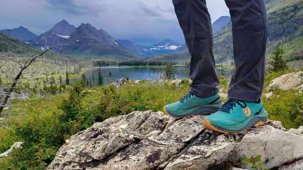 Knee-down view of a hiker standing on a white granite rock in a pair of teal Topo Athletic Ultraventure 3 hiking shoes in front a lake in Glacier National Park.