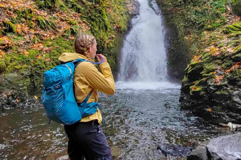 A hiker in front of a waterfall with the Gregory Juno 24 H2O hydration pack