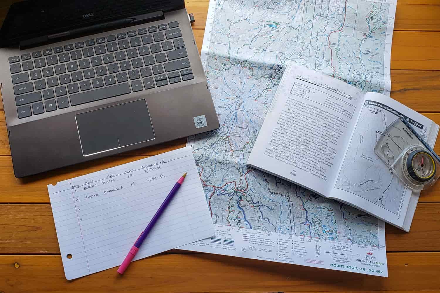 A desk with a laptop, map, compass, and handwritten notes 