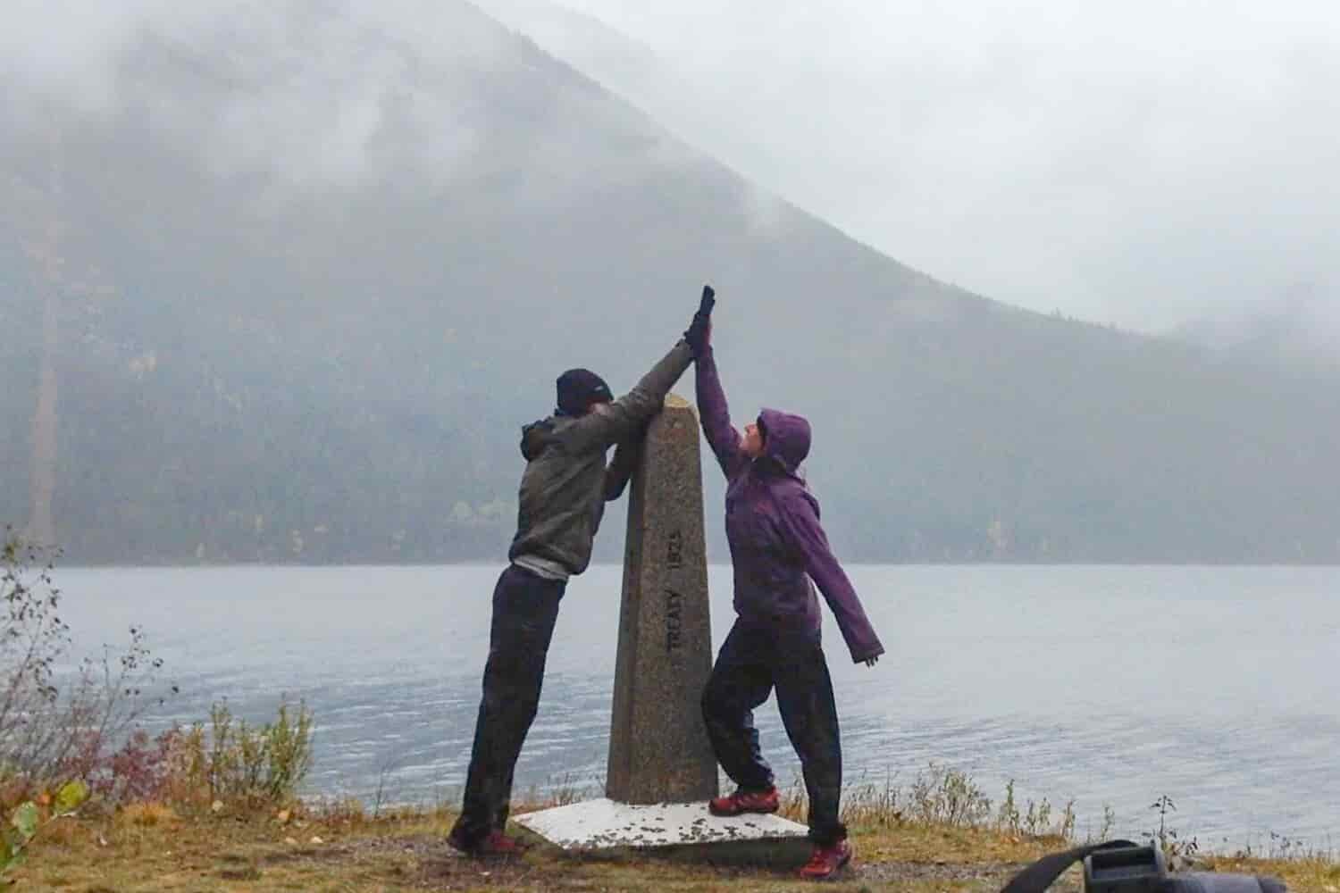 two backpackers high five in front of the CDT's northern terminus monument with a lake and mountain in the background in the rain.