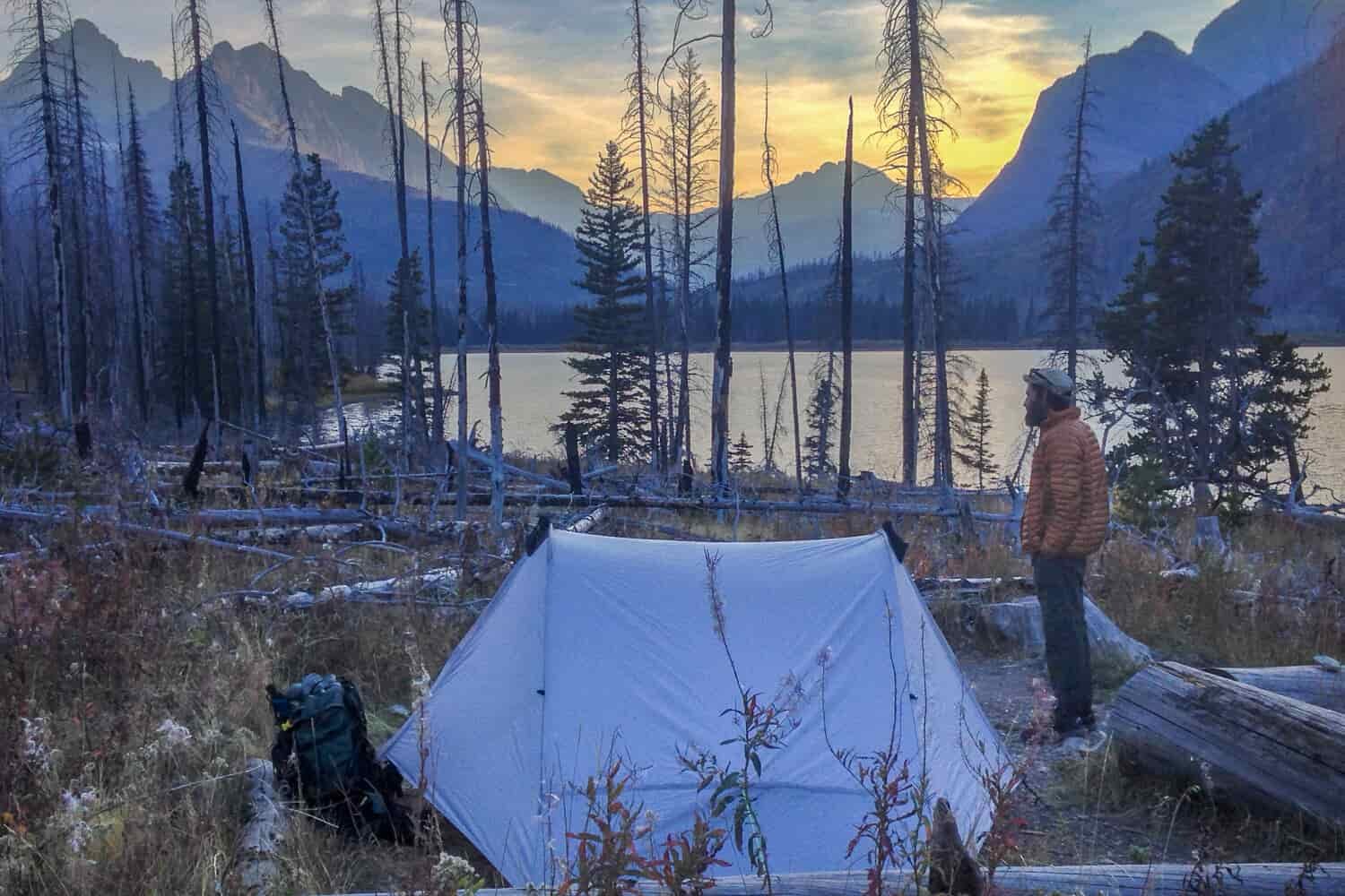 sunset behind mountains and a lake with a backpacker and his tent set up.