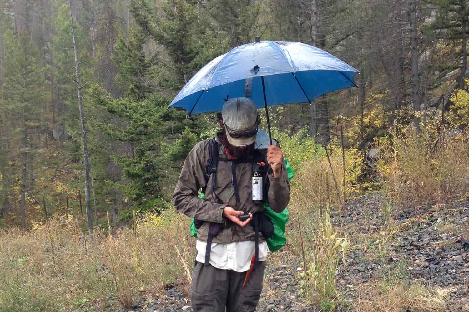 CDT thru-hiker using a GPS device to navigate while holding an umbrella and wearing bear spray