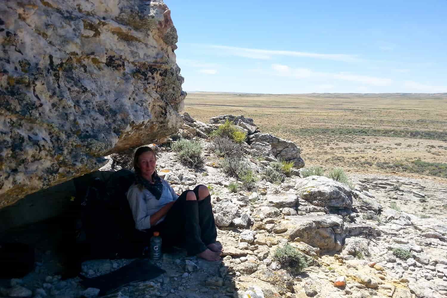 CDT thru-hiker sitting in the shade under a boulder with vast, flat, treeless plains in the background