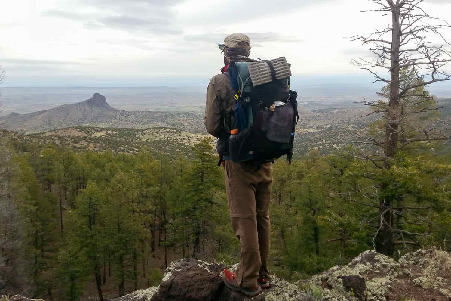 CDT thru-hiker with backpack on overlooking a ledge with trees and mountains below.