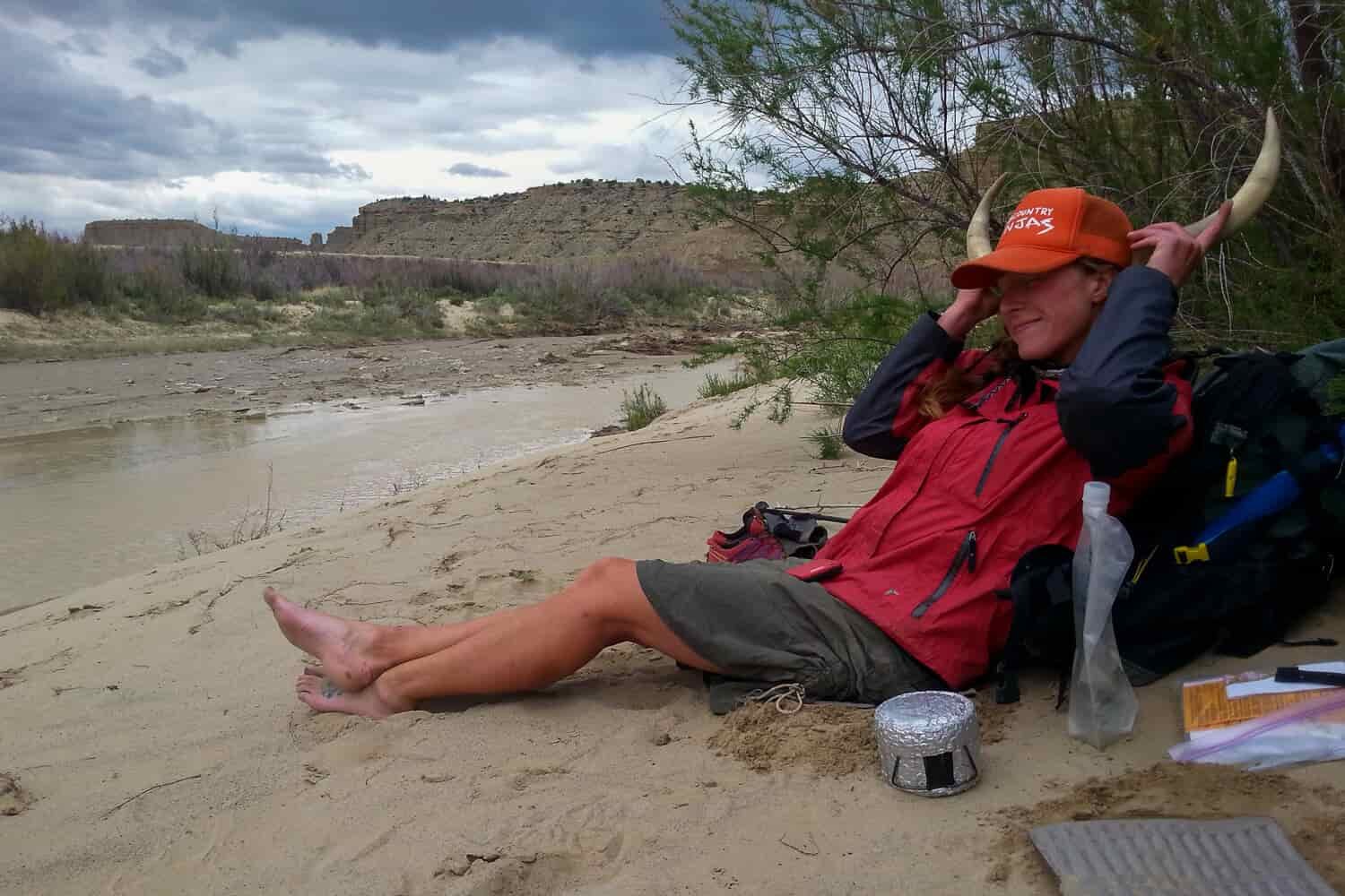 CDT thru-hiker holding up animal horns to their head while sitting alongside a river.