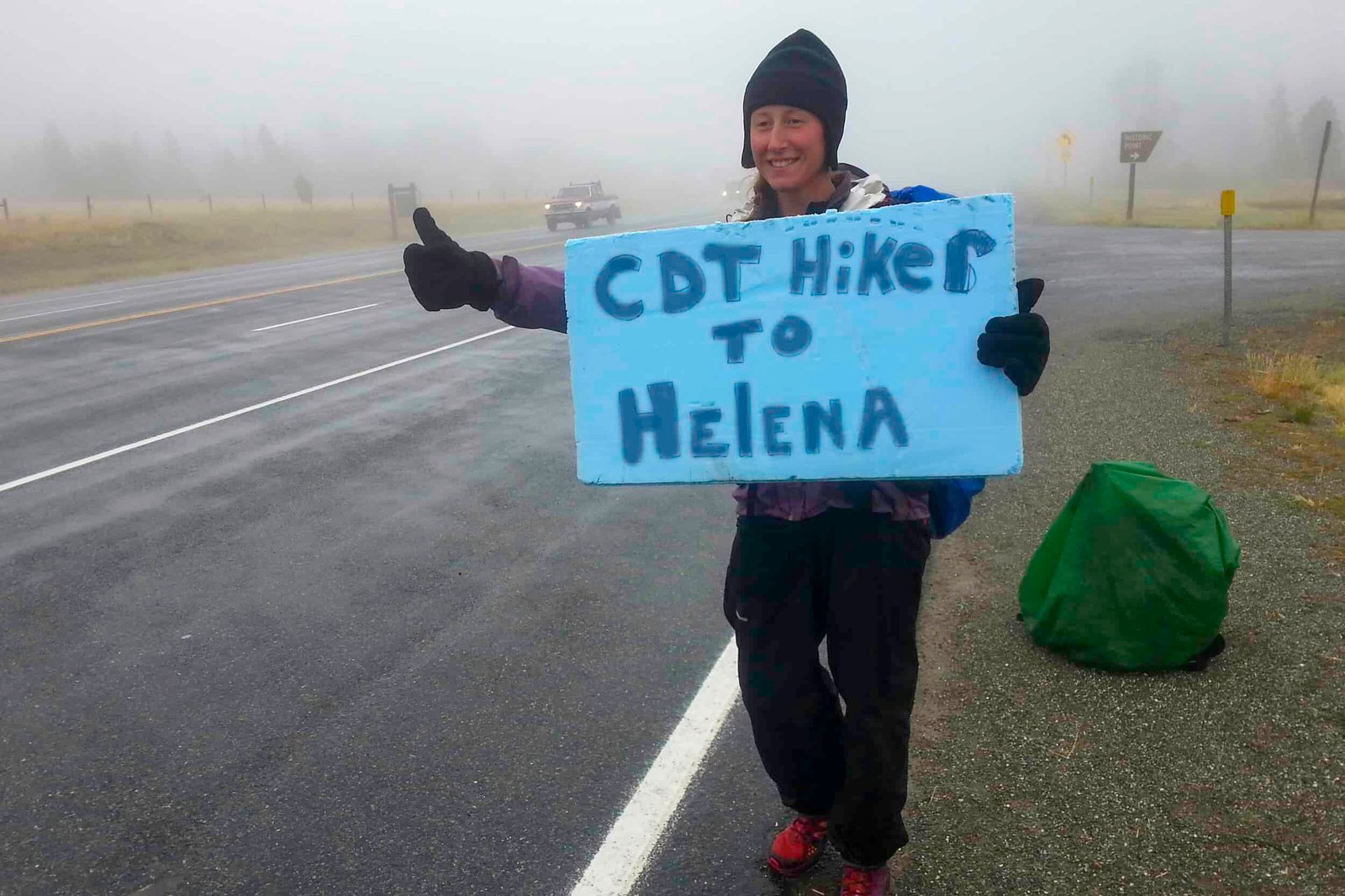 CDT thru-hiker holding a sign saying "CDT hiker to helena" and holding her thumb out to ask for a ride.