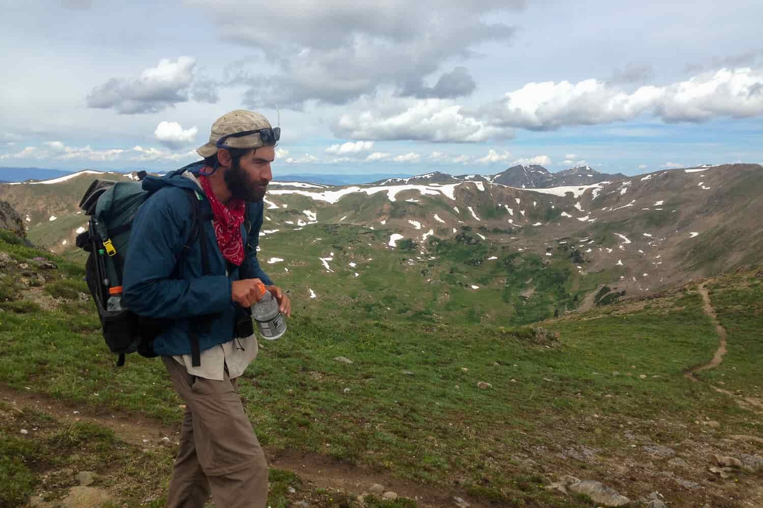 CDT thru-hiker backpacking on a high mountain pass with green and snow dotted peaks in the background