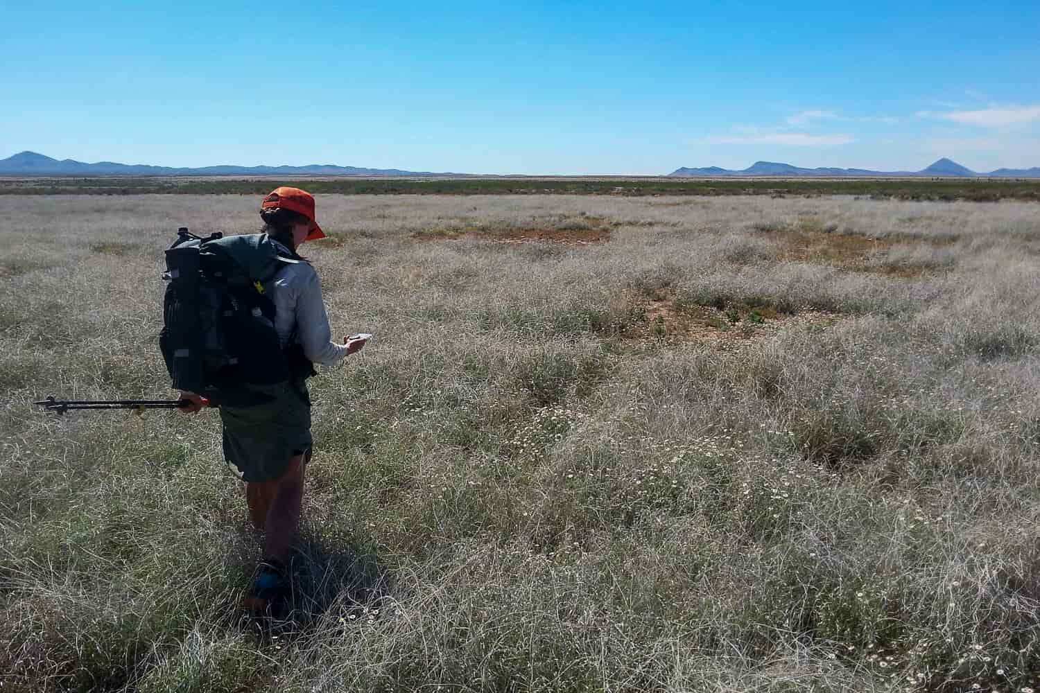 CDT thru-hiker holding a compass in a treeless, shrubby desert with mountains in the far off distance on a sunny day.