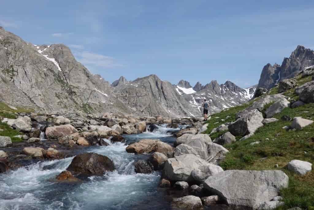 backpacker walks next to a rushing river below granite peaks in the summer in the wind river range