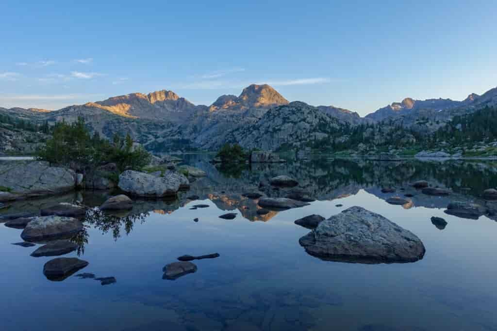a still lake with boulders and sunrise lighting up distant peaks