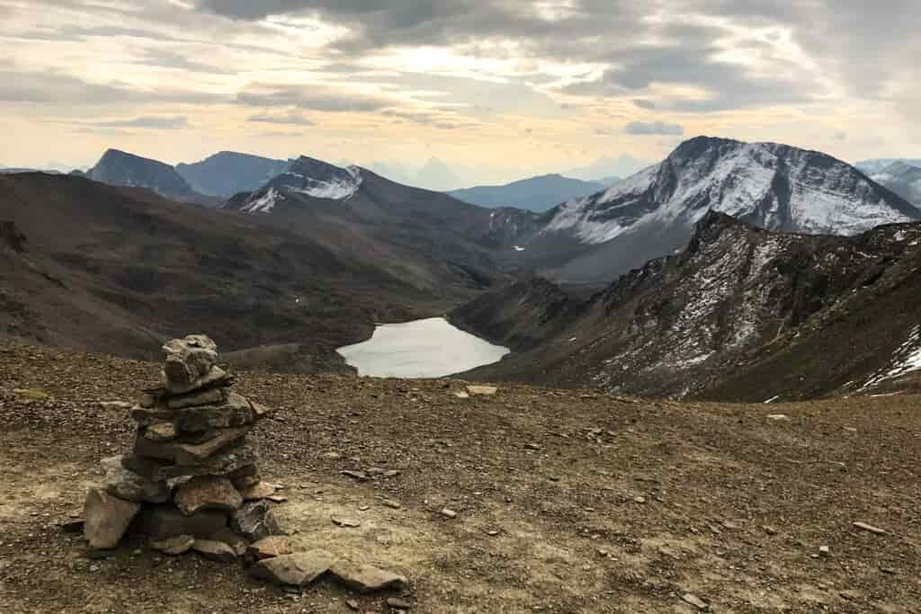 a rock cairn at a mountain pass overlooking a lake at sunset with snow dusted peaks surrounding the lake