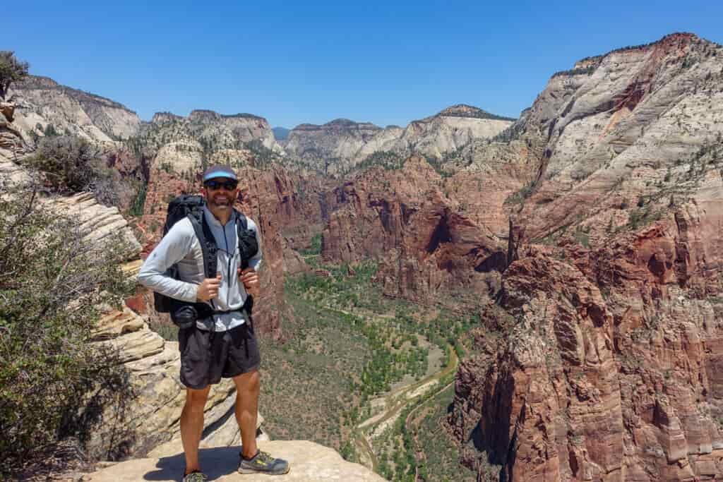 a backpacker stands on the top of a cliff to pose above zion canyon. You can see the road in the bottom of the green valley