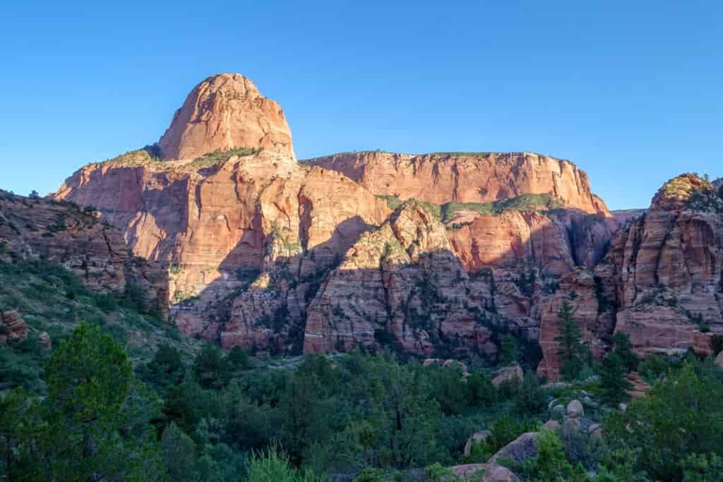 a mesa rising above a green valley floor lit up by the sun on a bright blue afternoon in zion national park on the trans-zion route