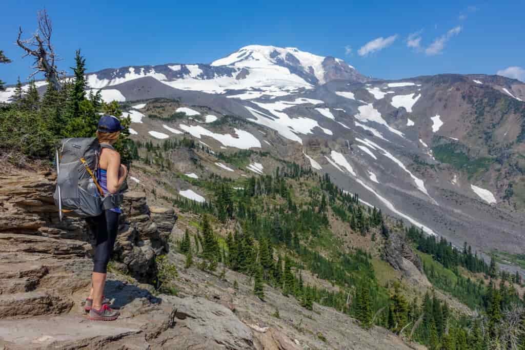 A female hiker stands at a ridge looking at snow-capped mountains in the distance 